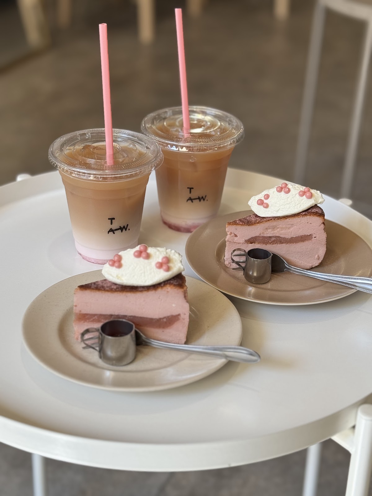 白いラウンドテーブルに2皿のさくらバスクチーズケーキとTAW.ロゴ入りアイスラテ2杯、ピンクストロー