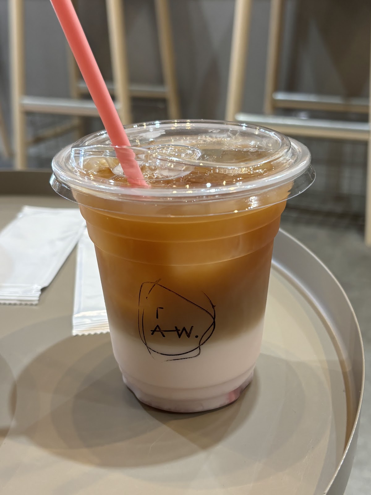 ステンレストレーの上に置かれたTAW.ロゴ入りアイスラテ。コーヒーと桜シロップの層が透明カップ越しに見える