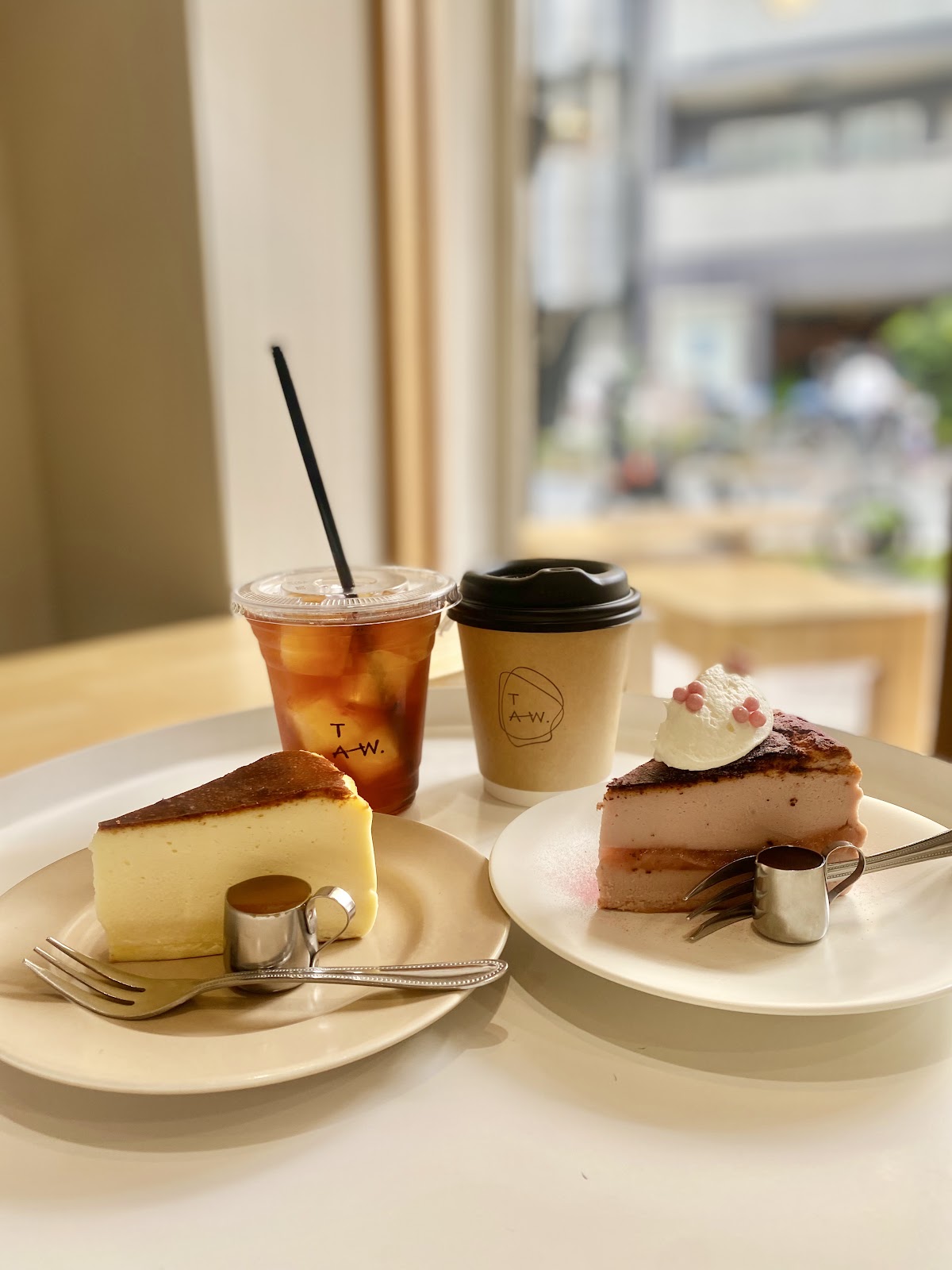 明るい窓際のテーブルにプレーンと桜フレーバー2種のバスクチーズケーキとTAW.ロゴ入りアイスコーヒー・ホットコーヒーカップ
