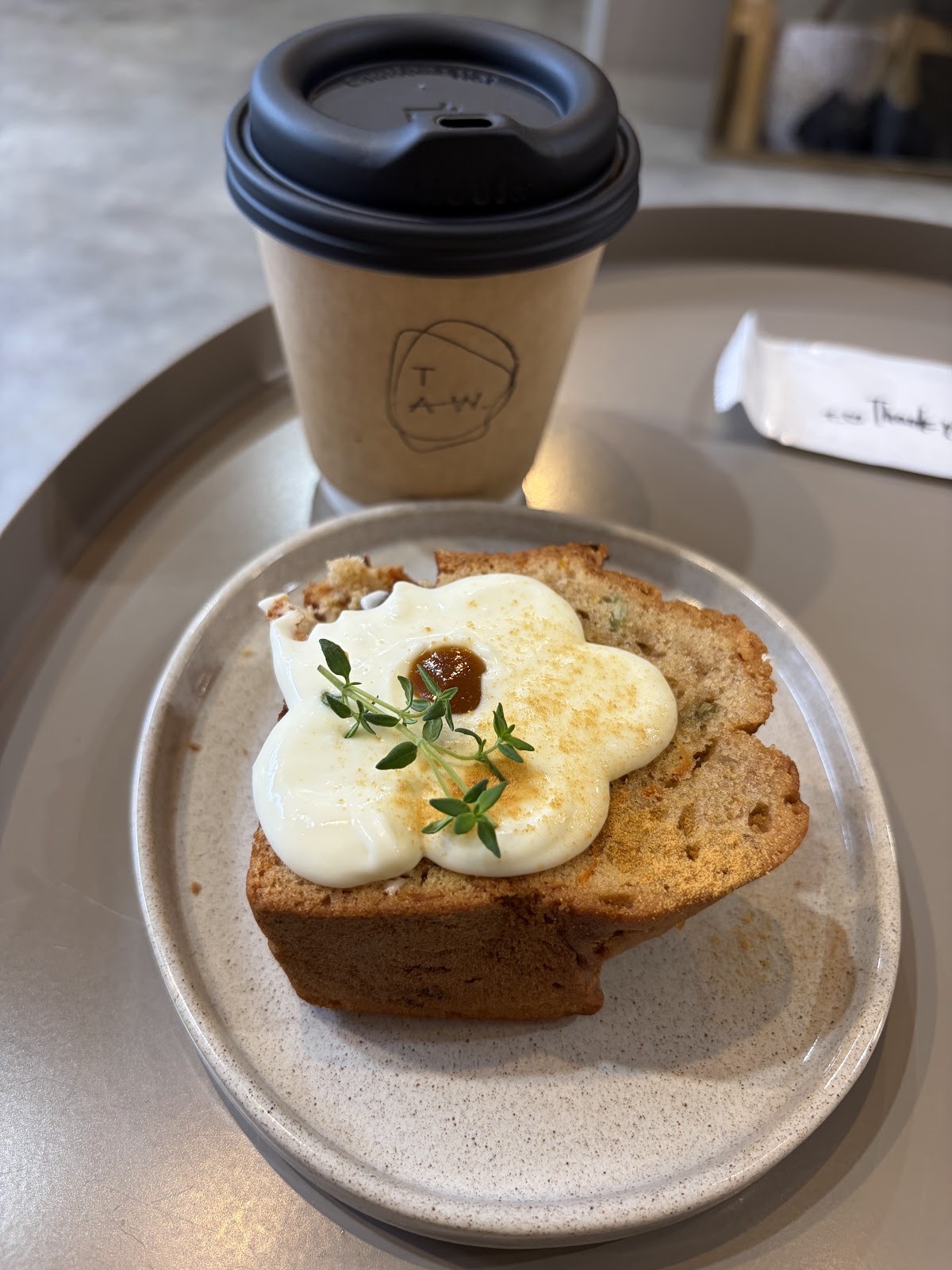丸いステンレストレーの上にTAW.ロゴ入りカップとキャロットケーキ。花形クリームとハーブが乗った厚切りの焼き菓子